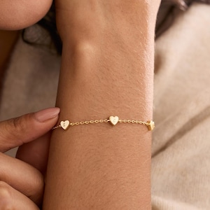 Könnte beinhalten: Ein zartes Goldkettenarmband mit drei kleinen, gleichmäßig verteilten herzförmigen Anhängern. Jeder herzförmige Anhänger ist mit einem einzelnen Buchstaben graviert, namentlich den Buchstaben S und C, verbunden durch eine feine Goldgliederkette.