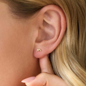 May include: A small gold stud earring shaped like the lowercase letter m. The earring is positioned on the earlobe, featuring a polished metallic finish and a simple, delicate script font design that sits flush against the skin of the ear.