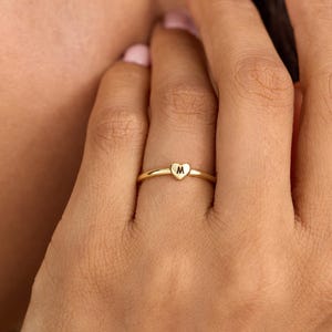 Peut inclure: Une fine bague dorée ornée d'un petit cœur poli centré sur l'anneau. Le cœur est gravé de la lettre majuscule M dans une police simple, sombre et sans empattement. La bague présente une finition métallique lisse et réfléchissante.