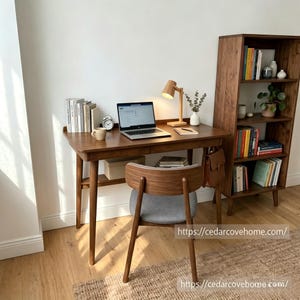 Op de afbeelding: Een bureau van donker hout met twee lades en taps toelopende poten, gecombineerd met een bijpassende stoel met een gebogen rugleuning en grijze bekleding. Op het bureau staan een laptop, een houten bureaulamp, een mok, boeken en een klok.
