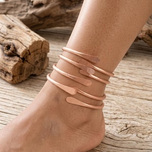 May include: A copper-colored metal anklet featuring a multi-coil wrap design. The anklet consists of a single continuous wire that spirals around the ankle, ending in flattened, rounded tips. The metal has a smooth, polished finish.