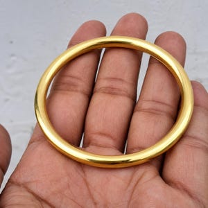 May include: A smooth, polished gold-tone bangle bracelet with a simple, continuous circular design. The metal surface is highly reflective, showing a bright yellow-gold color throughout the entire circumference of the solid, rounded band.
