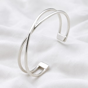 Può includere: Un bracciale rigido color argento lucido dal design aperto ed essenziale, composto da due fasce che si incrociano sulla parte anteriore. Il bracciale presenta una superficie liscia e riflettente, terminando con due terminali piatti e rettangolari.