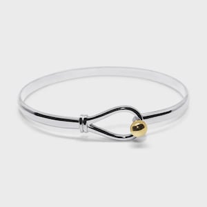 May include: A silver-toned bangle bracelet featuring a smooth, polished finish. The design includes a central loop closure mechanism that secures with a small, spherical gold-toned bead at the end, creating a clean and simple circular silhouette.