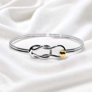 Puede incluir: Una pulsera rígida de tono plateado que presenta un diseño de nudo central estilo infinito. La pulsera está construida con una banda de metal pulido y liso, e incluye una pequeña cuenta esférica de acento en tono dorado en un lado del nudo.