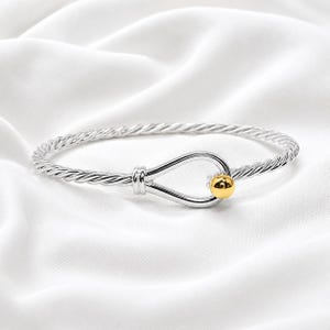 Puede incluir: Un brazalete de cable trenzado en tono plateado con un cierre central en forma de bucle de gota. El cierre está acentuado con una sola cuenta esférica pulida en tono dorado en el extremo del bucle, creando un contraste distintivo de color y textura.
