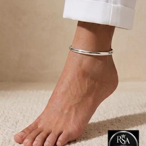 Puede incluir: Una tobillera de plata lisa y pulida que presenta un diseño de banda circular simple y continua. La tobillera se lleva alrededor del tobillo, descansando justo encima del pie, y tiene un acabado metálico reflectante y uniforme.