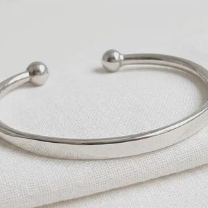 May include: A polished silver-tone metal cuff bracelet featuring a smooth, curved band that tapers slightly toward the ends. Each terminal of the open-ended bracelet is finished with a small, rounded spherical ball detail, creating a clean design.