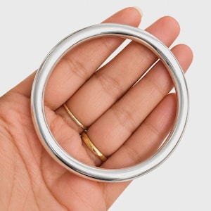 May include: A smooth, polished silver-tone bangle bracelet with a simple, circular design. The bangle features a continuous, rounded profile with a reflective metallic finish, showcasing a clean and uniform appearance throughout the entire piece.