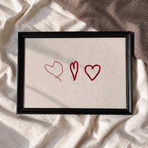 Puede incluir: Un marco rectangular negro que contiene un panel de tela beige claro con tres contornos de corazones rojos bordados a mano. Los corazones están dispuestos horizontalmente, cada uno con un patrón de puntada de estilo dibujado a mano.