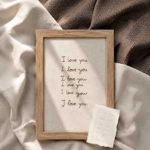 Op de afbeelding: Een rechthoekige houten lijst met een lichtgekleurd stoffen paneel waarop de zin I love you zes keer is geborduurd met donker garen. Daaronder ligt een klein, gescheurd stukje wit papier met dezelfde zin geschreven in lichte inkt.