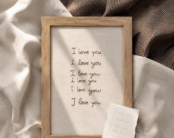 May include: A rectangular wooden frame containing a light-colored fabric panel embroidered with the phrase I love you repeated six times in dark thread. A small, torn piece of white paper with the same phrase written in light ink rests below.