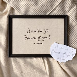 Può includere: Una cornice rettangolare nera contenente un inserto in tessuto beige con il testo nero ricamato "I am So Proud of you! x mom" e un simbolo a forma di cuore. Un piccolo pezzo di carta bianca strappata con lo stesso messaggio scritto a mano è appoggiato sulla cornice.
