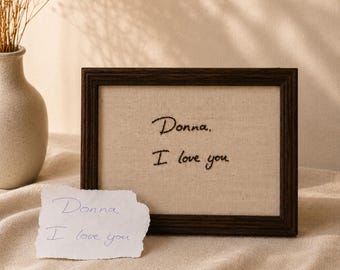 May include: A dark brown rectangular wooden frame containing a beige fabric insert with the black embroidered text "Donna, I love you". In front of the frame sits a small, torn piece of white paper with the same text written in blue ink.