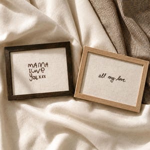 Puede incluir: Dos cuadros rectangulares con fondo de tela color hueso y texto bordado en negro. El marco izquierdo es de madera oscura con el texto MAMA I love You xxx. El marco derecho es de madera clara con el texto all my love.