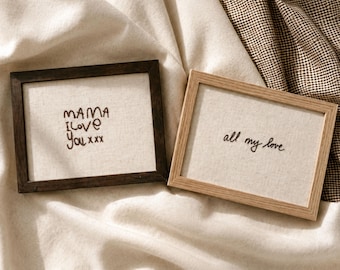 May include: Two rectangular framed wall art pieces featuring off-white fabric backgrounds with black embroidered text. The left frame is dark wood with the text MAMA I love You xxx. The right frame is light wood with the text all my love.