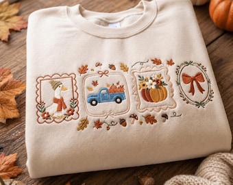 May include: A beige crewneck sweatshirt featuring embroidered autumn-themed designs. The embroidery includes a goose wearing a scarf, a blue pickup truck carrying pumpkins, a pumpkin floral arrangement, and a bow, all framed in decorative borders.