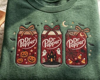 May include: A forest green crewneck sweatshirt featuring an embroidered design of three Dr Pepper soda cans. Each can is topped with a bow and decorated with Halloween motifs including pumpkins, ghosts, a haunted house, spiders, and candy corn.