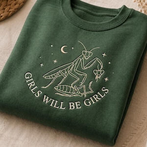 Puede incluir: Una sudadera de cuello redondo en color verde bosque oscuro con un bordado de dos mantis religiosas. El dise&ntilde;o incluye una luna creciente, estrellas y el texto "GIRLS WILL BE GIRLS" en hilo claro centrado en el pecho.