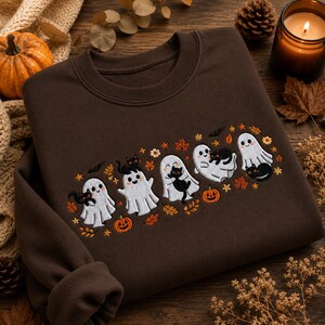 May include: A dark brown crewneck sweatshirt featuring an embroidered design of five white ghosts, each holding a small black cat. The embroidery includes orange pumpkins, small bats, and scattered autumn leaves in shades of orange and yellow.