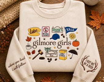 May include: A beige crewneck sweatshirt featuring embroidered icons and text related to Gilmore Girls. The design includes Luke's sign, a no cell phones sign, Doose's Market sign, a Yale flag, a blue cap, a pumpkin, a gavel, and a Dragonfly Inn sign.
