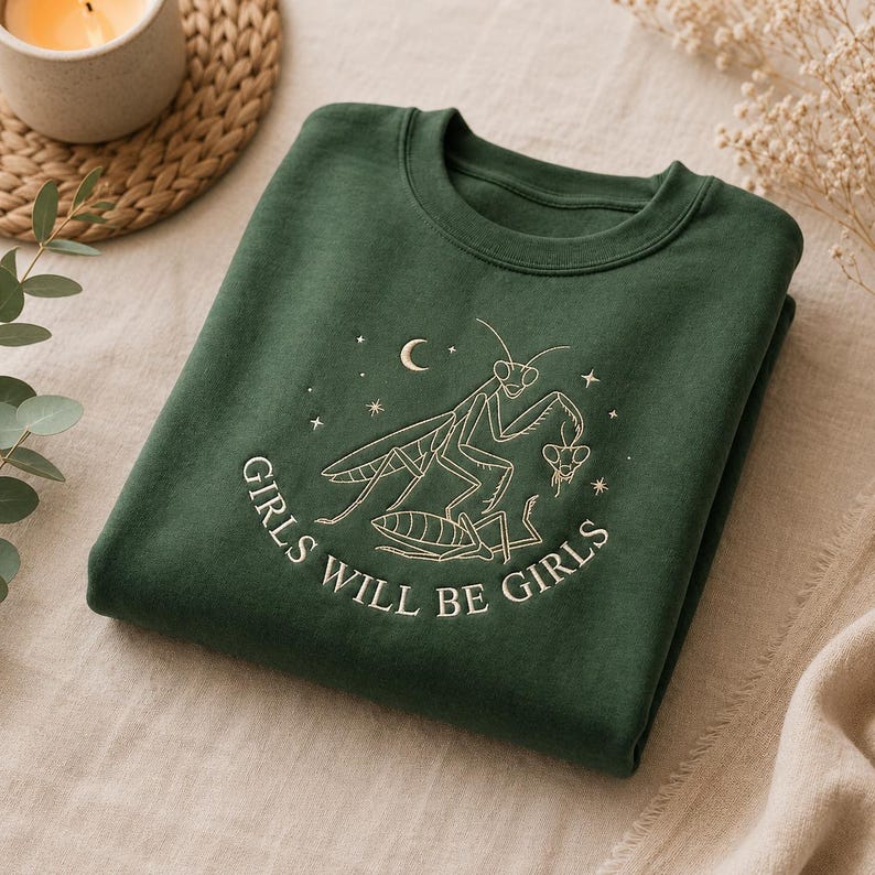 Puede incluir: Una sudadera de cuello redondo en color verde bosque oscuro con un bordado de dos mantis religiosas. El dise&ntilde;o incluye una luna creciente, estrellas y el texto "GIRLS WILL BE GIRLS" en hilo claro centrado en el pecho.