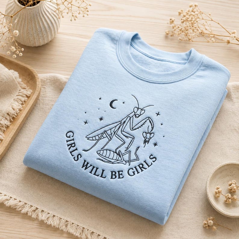 Puede incluir: Una sudadera azul claro de cuello redondo con bordado negro de dos mantis religiosas. Una mantis est&aacute; sobre la otra, rodeada de estrellas y una luna creciente. El texto GIRLS WILL BE GIRLS est&aacute; bordado en un arco debajo de los insectos.