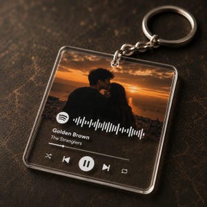 May include: A square clear acrylic keyring featuring a printed sunset photograph of two people. The design includes a white Spotify-style music player interface with the text Golden Brown by The Stranglers, a progress bar, and playback icons.