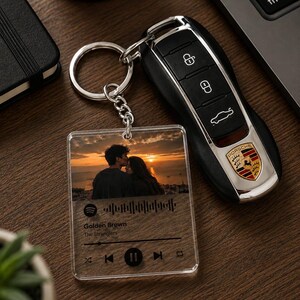 May include: A clear acrylic square keyring featuring a printed photograph of a couple at sunset, a Spotify-style waveform, and the text Golden Brown by The Stranglers, attached to a silver metal ring next to a black Porsche car key fob.