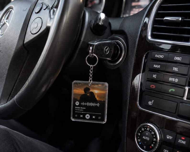 May include: A square, clear acrylic keyring hanging from a car ignition key. The keyring features a printed image of a couple at sunset, a Spotify-style waveform, and the text Golden Brown by The Stranglers with playback control icons.
