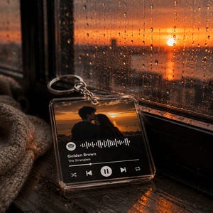 May include: A square clear acrylic keyring featuring a printed music player interface. The design displays a sunset silhouette of a couple, a Spotify code, and the text Golden Brown by The Stranglers with standard playback control icons.