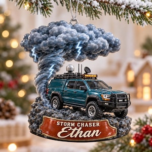Op de afbeelding: Een hangend ornament met een blauwgroene pick-up truck uitgerust met stormjagersuitrusting, onder een grote, wervelende grijze tornado met blauwe bliksemschichten. Een bruine plaquette aan de basis toont de tekst "STORM CHASER Ethan".