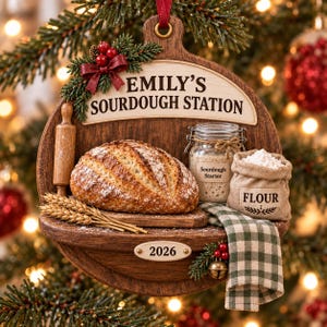 May include: A wooden Christmas ornament shaped like a cutting board with the text EMILY'S SOURDOUGH STATION. It features a loaf of bread, a rolling pin, a jar labelled Sourdough Starter, a sack labelled FLOUR, wheat stalks, and the year 2026.