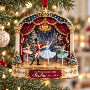 May include: A decorative Christmas ornament featuring a Nutcracker stage scene with a Nutcracker, Clara, and ballet dancers. The base displays the text THE NUTCRACKER 2026 Sophia AS CLARA. The scene includes red curtains, a chandelier, and a mouse king.