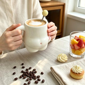 Può includere: Una tazza in ceramica bianca con una forma arrotondata simile a un corpo e un piccolo punto rosa, riempita di caffè. Un cucchiaino in metallo con un'estremità sferica giallo pallido riposa all'interno. Chicchi di caffè scuro sono sparsi vicino.