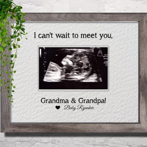 May include: A framed ultrasound image on white textured paper with the black text I can't wait to meet you, Grandma & Grandpa! followed by a heart symbol and the text Baby Rynaker. The frame is a dark gray wood with a visible grain pattern.