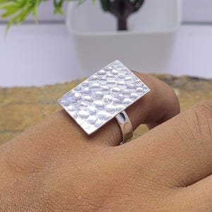 May include: A silver-toned ring featuring a large, flat, rectangular face with a textured, hammered surface pattern. The band of the ring is smooth and metallic, connecting to the underside of the rectangular plate. The design is simple and bold.
