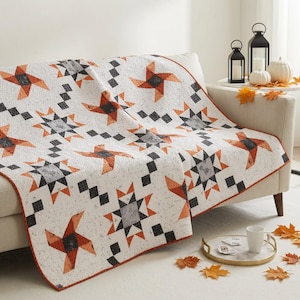 May include: A quilt featuring a repeating pattern of orange and grey star-like shapes and black diamond accents on a white background. The fabric has a subtle speckled texture throughout, and the edges are finished with a thin orange binding.