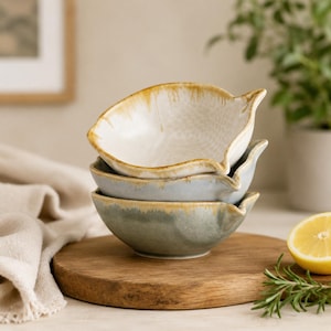 May include: A stack of three ceramic bowls with a leaf-like shape and a small pouring spout. The top bowl is cream with a golden-brown rim, the middle is light blue, and the bottom bowl is a muted sage green, each featuring a reactive glaze finish.