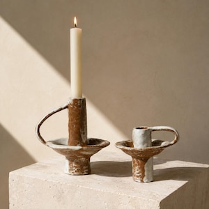 May include: Two ceramic candle holders with a rustic, textured finish in shades of brown and white. Each holder features a wide, shallow base, a central cylindrical column, and a curved handle. A single tall, cream-colored taper candle is lit.
