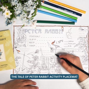 May include: A white paper activity placemat featuring Peter Rabbit line art, word scrambles, and an I Spy game. The sheet includes illustrations of garden tools, vegetables, and birds. A hand holds a black pencil, actively filling in the page.