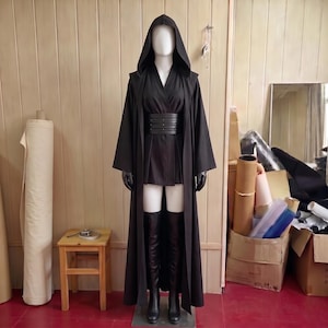 以下が含まれることがあります： 黒いフード付きローブ、黒いチュニック、銀色のスタッズが付いた幅広の多層ベルト。黒い手袋と膝上丈の黒いブーツを合わせた衣装が、明るい木目調の壁を背景にしたマネキンに着用されています。