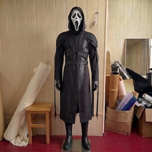Pode incluir: Um traje de corpo inteiro com uma máscara branca de Ghostface com capuz preto, um casaco longo preto com painéis estruturados nos ombros, tiras pretas integradas com fivelas no peito e na cintura, mangas compridas e botas pretas.