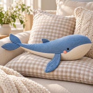 May include: A whale-shaped plush toy featuring a blue textured upper body and a cream-colored underbelly. The toy has a small black eye, a round pink cheek detail, and visible white stitching along the fins, tail, and body seams.