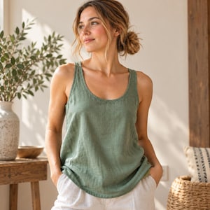May include: A sage green sleeveless vest top with a scoop neckline and a relaxed fit. The garment features a textured fabric appearance and is worn with light-coloured trousers. The design is simple, featuring wide shoulder straps and a curved hemline.