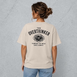 Puede incluir: Una camiseta beige de manga corta con texto negro en la espalda que dice THE OVERTHINKER, un gráfico de garabato circular debajo y el texto THINKING TOO MUCH SINCE ALWAYS debajo del gráfico en una fuente sans-serif simple.