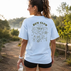 May include: A light blue t-shirt featuring a dark green graphic on the back with the text "RUN CLUB" and "RUN MORE WORRY LESS." The design includes illustrations of running shoes, a cap, a water bottle, a banana, a race bib, and other gear.
