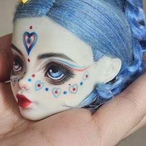May include: A doll head with bright blue hair styled in a high ponytail and a small yellow star accessory. The face features pale skin, painted blue and red heart motifs on the forehead and cheeks, dark eyelashes, blue irises, and bright red lips.