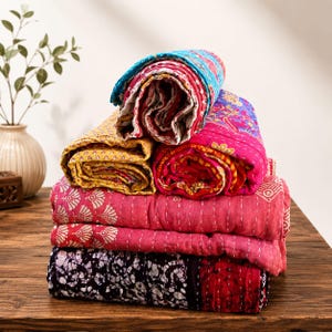 May include: A stack of five folded cotton quilts featuring various patterns and colors, including bright pink, yellow, blue, and a black and white batik print. Each quilt displays visible hand-stitched kantha embroidery with intricate running stitches.