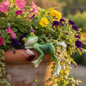 Puede incluir: Una figura de rana verde con ojos amarillos descansando en el borde de una maceta de terracota, sosteniendo un pequeño vaso transparente lleno de líquido rojo oscuro, rodeada por un arreglo vibrante de flores rosas, amarillas y moradas.
